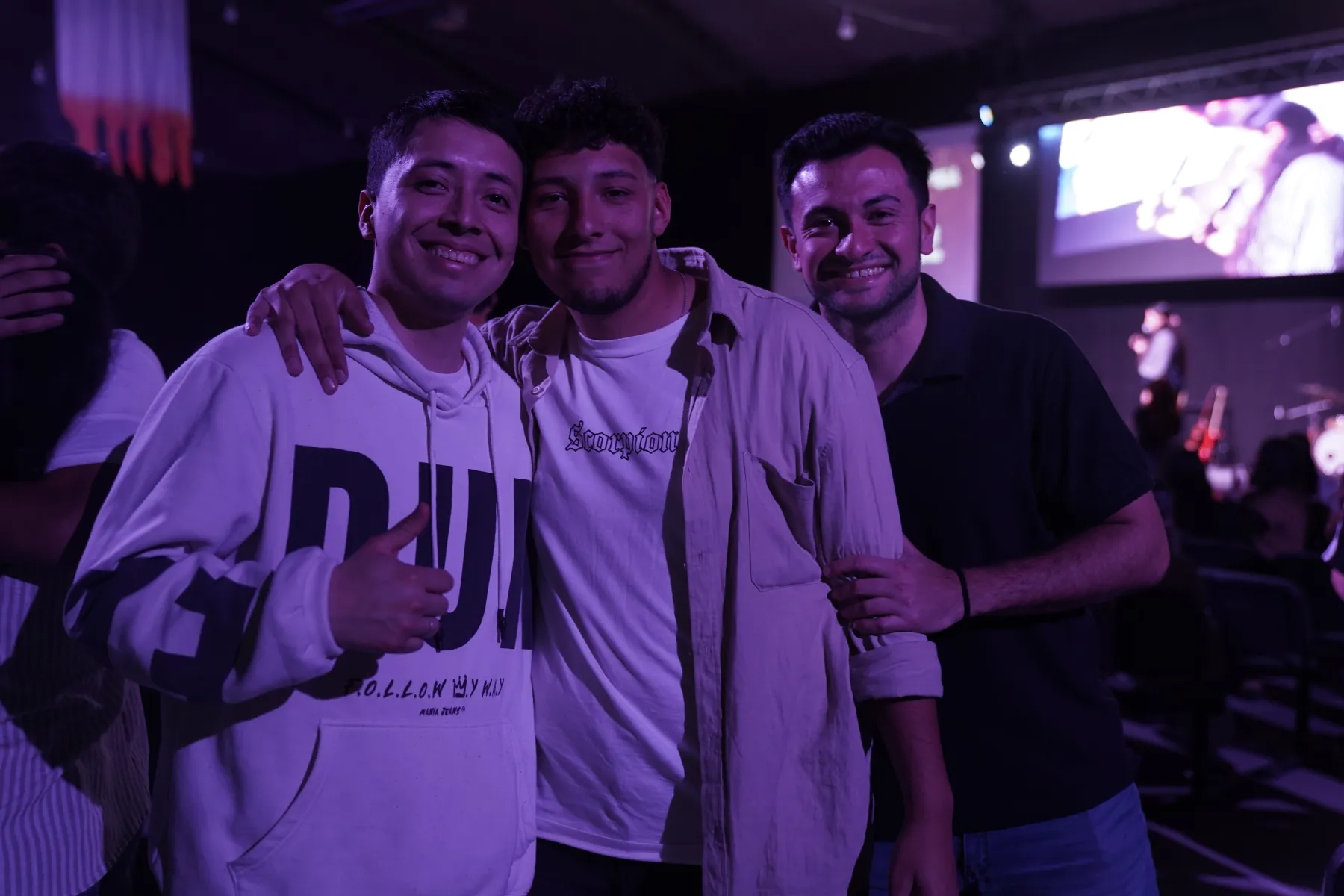 Tres chicos abrazados y sonriendo, con el escenario iluminado detrás.